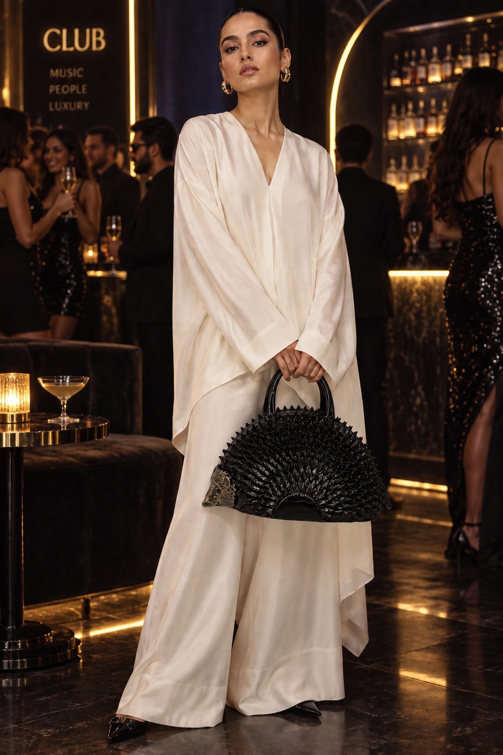 A woman in a luxurious club setting wearing a flowing white satin robe and holding a black spiky handbag.