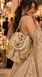A woman in a heavily embellished beige and gold traditional outfit carries a large ornate gold shoulder bag decorated with pearls, beads, and tassels in a festive indoor setting with warm lights and blurred people in the background.