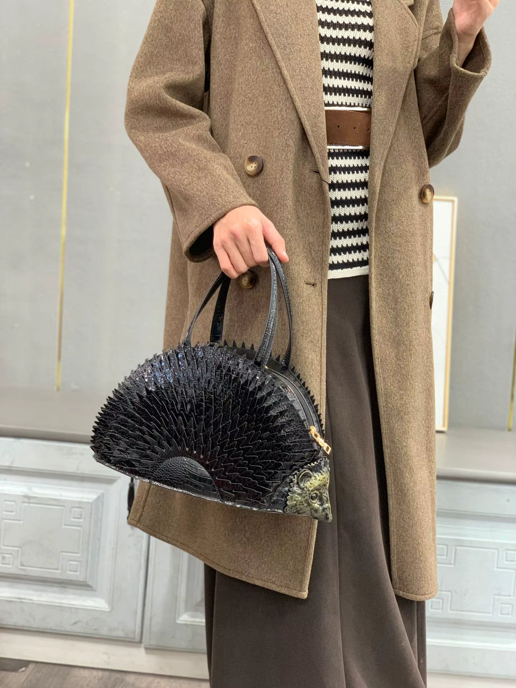 Person wearing a brown coat holding a black handbag in front of a mirror.