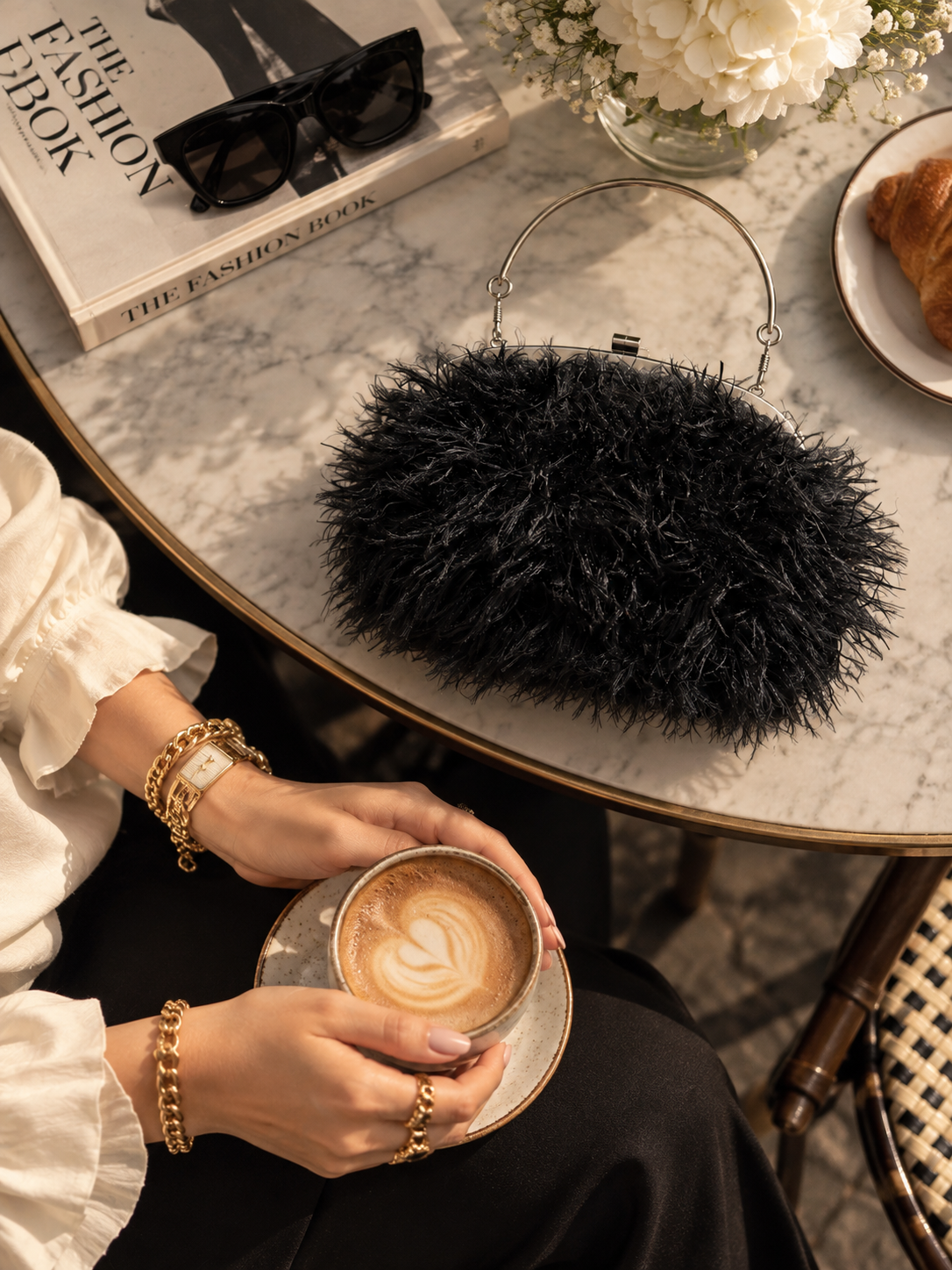 A black feathered clutch purse with a metal frame and top handle sits on a marble cafe table next to a person holding a latte. The scene includes sunglasses on a book, white flowers, and a croissant, creating a stylish lifestyle setting. clutch by capybaraluxe.com