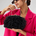 A woman in a bright pink blazer holds a black feathered handbag with a gold-tone metal handle and frame against a light gray studio background.