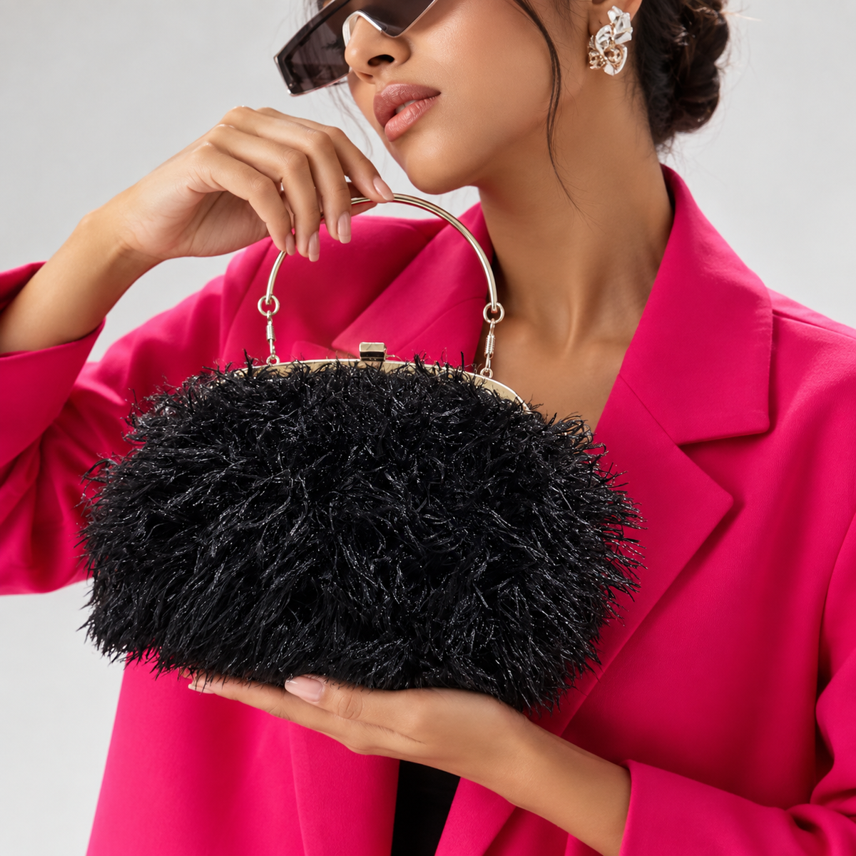 A woman in a bright pink blazer holds a black feathered handbag with a gold-tone metal handle and frame against a light gray studio background.