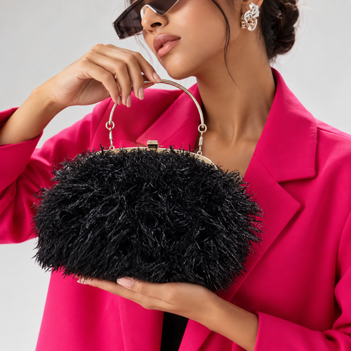 A woman in a bright pink blazer holds a black feathered handbag with a gold-tone metal handle and frame against a light gray studio background.