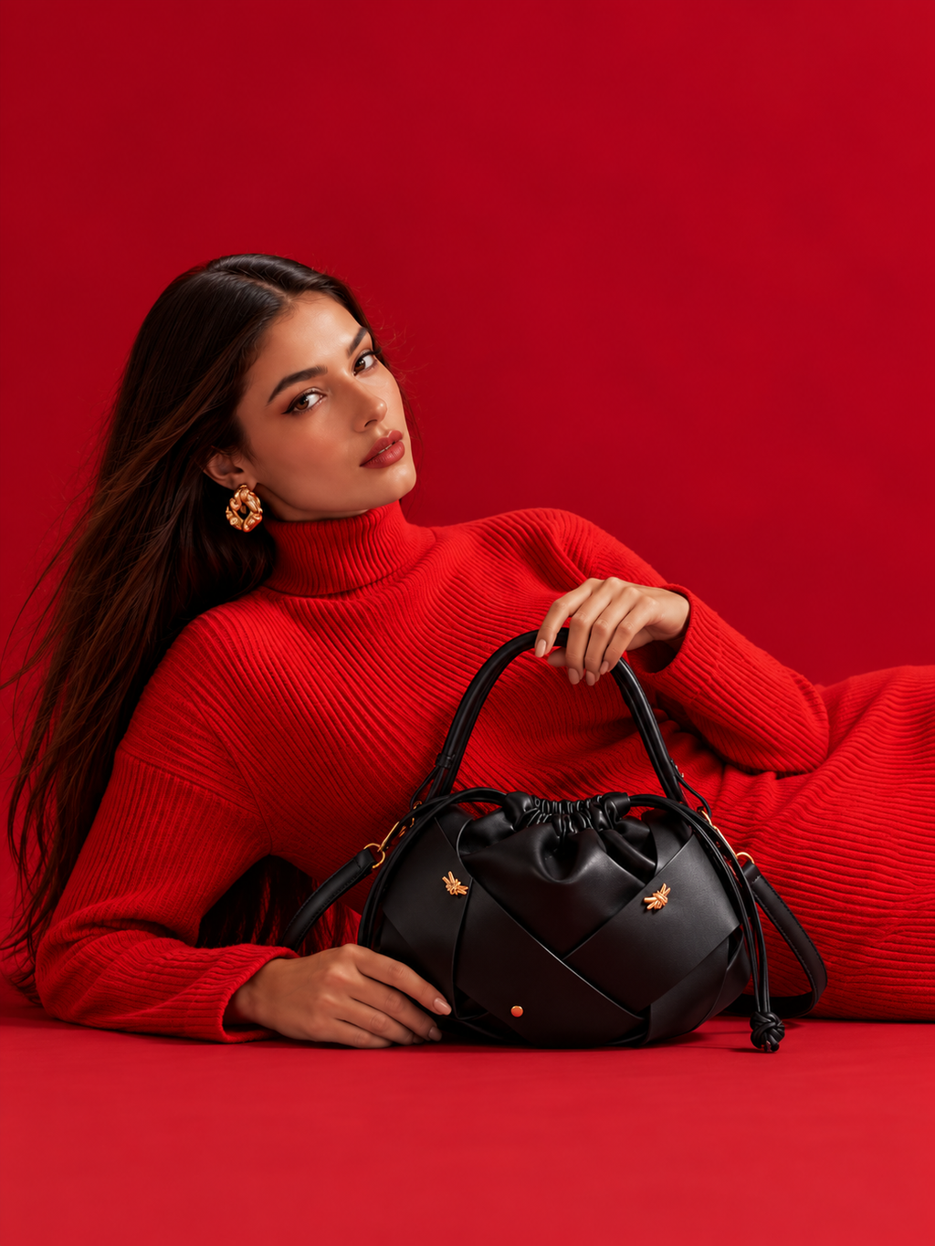 A woman in a bright red ribbed turtleneck sweater reclines on a solid red studio background, holding a black leather handbag with gold-tone hardware and small bee motifs.