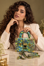 A woman in a white blazer holds a structured handbag with a vibrant green, blue, and yellow reptile-embossed pattern. The matching shoulder strap rests on the marble table in front of her.